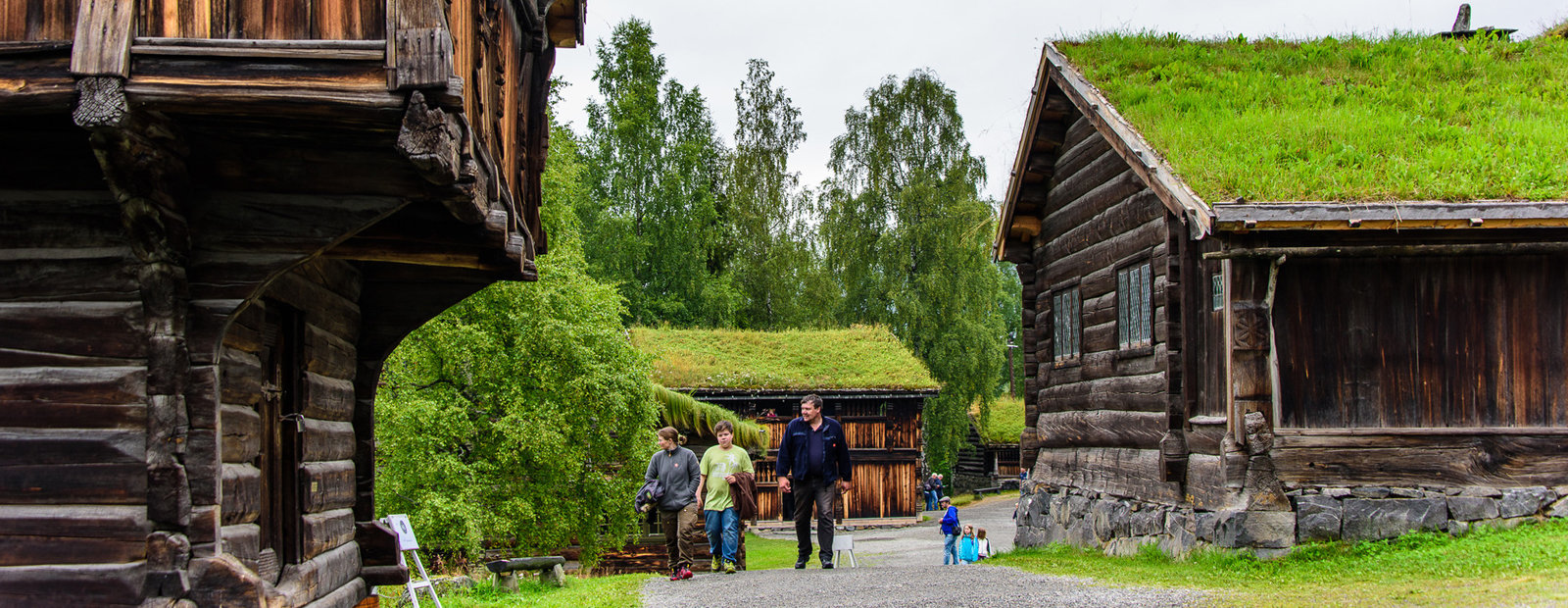  Folk går på grusvei og titter på gamle tømmerhus på friluftsmuseet Maihaugen i Lillehammer. 